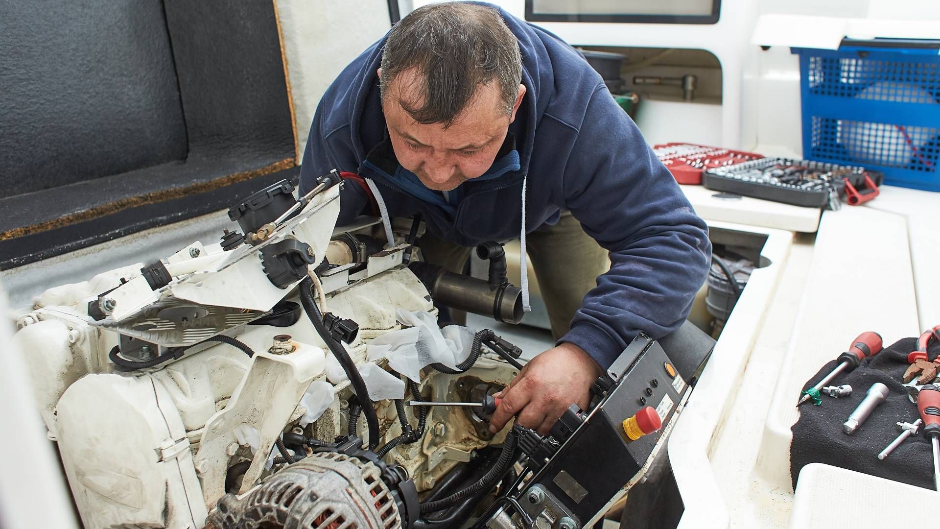 Man in blue sweatshirt working on a boat engine. White boat interior, tools visible.
