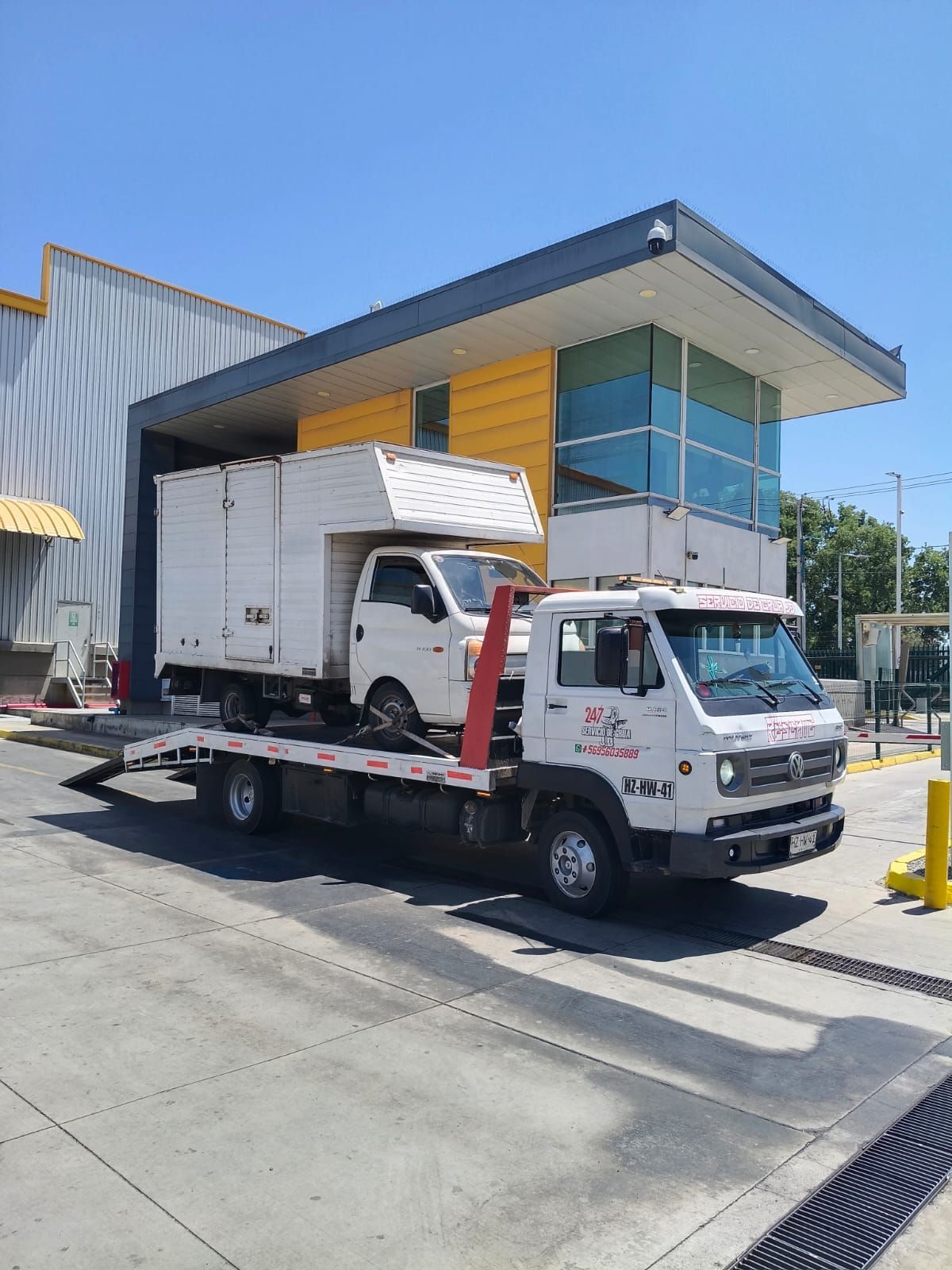 Una grúa de plataforma blanca transportando un camión de caja blanca más pequeño estacionado frente a un edificio moderno.