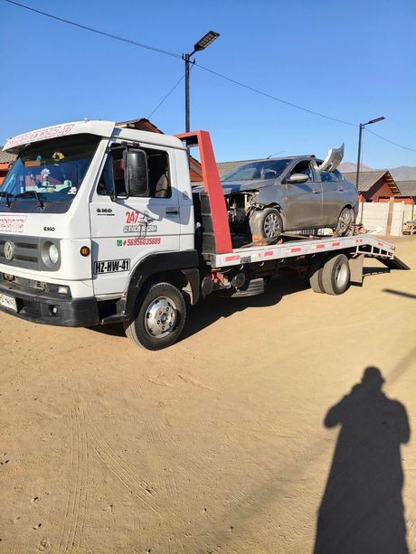 Una grúa blanca transporta un sedán gris dañado por un camino de tierra bajo un cielo azul despejado.