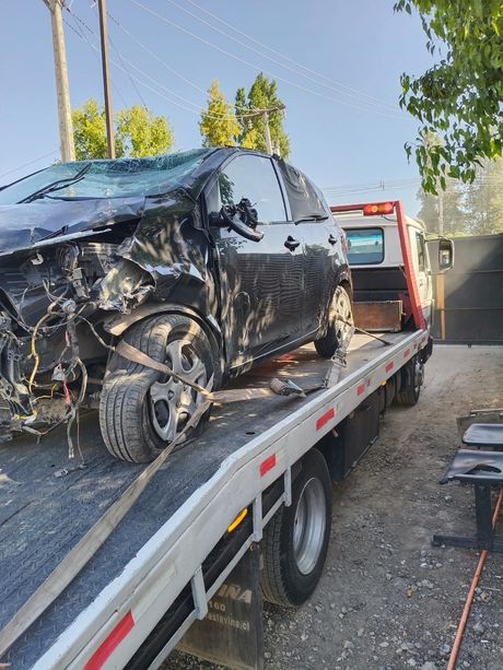 Un coche negro gravemente dañado se encuentra asegurado sobre la plataforma de una grúa en un entorno al aire libre.