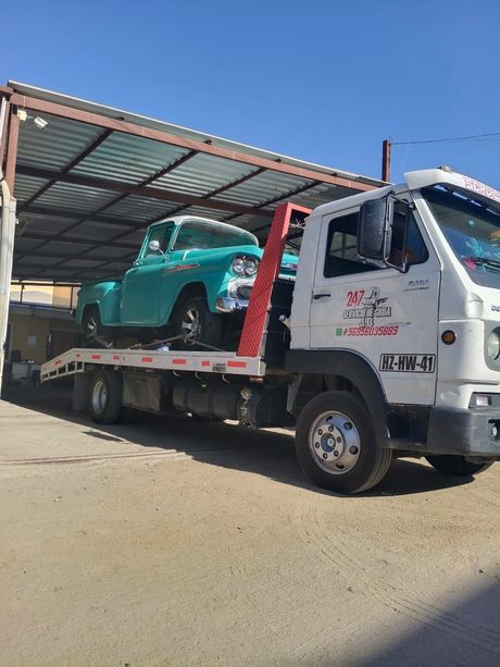 Una camioneta pickup antigua de color verde azulado cargada sobre una grúa de plataforma blanca bajo una cubierta metálica.