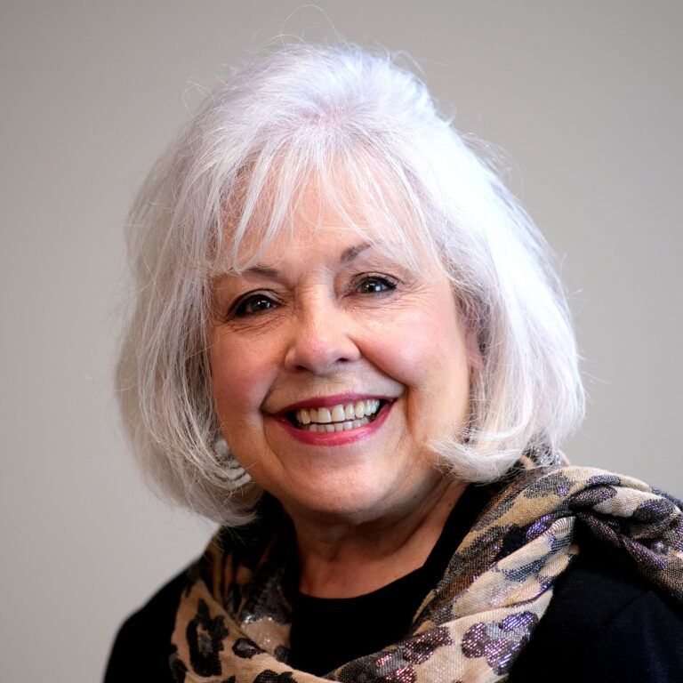 A woman with white hair is wearing a scarf and smiling.