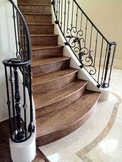A curved wooden staircase with ornate black wrought-iron railings, beginning on a marble floor in a bright interior space.