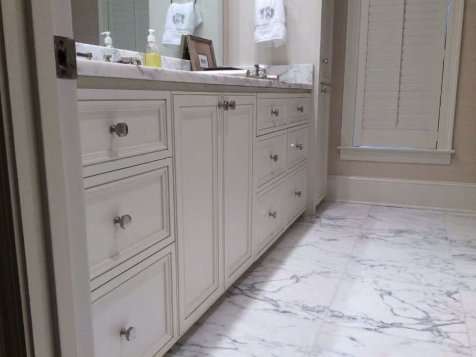 A white vanity with drawers and cabinets sits on a white marble-tiled floor in a bathroom with shuttered windows.