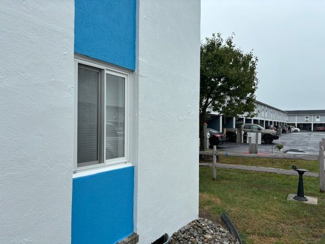 A white building with blue trim, a window, and a grassy lawn on an overcast day.