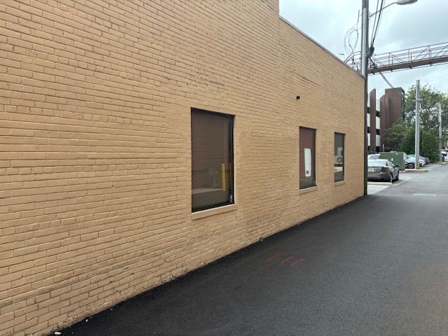 Tan brick building with three windows, asphalt road, parked cars, overcast sky.