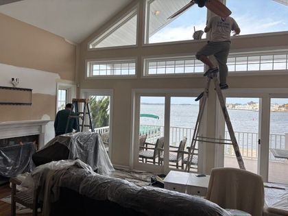 Two painters work inside a living room, one on a ladder, with a view of the water outside.