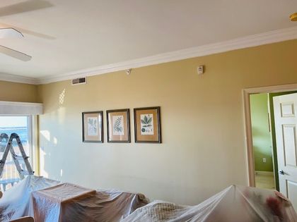 Beige living room with crown molding, three framed pictures, couch covered with plastic, and a doorway.