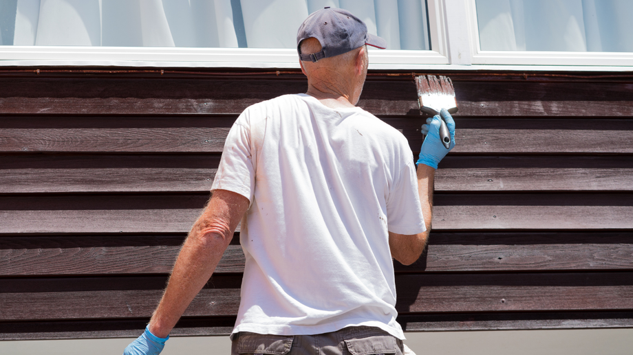 Man in a white shirt and blue gloves painting a brown wooden wall with a brush near a white window.