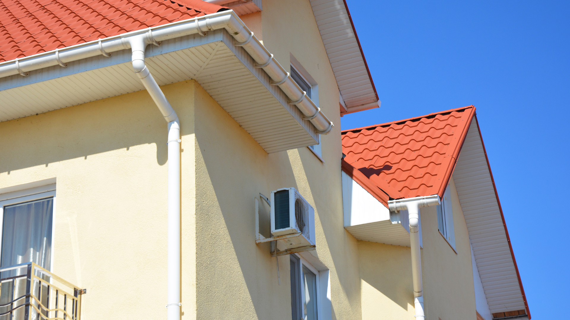Two-story house with red roof, yellow siding, white trim, gutters, and AC unit against a blue sky.