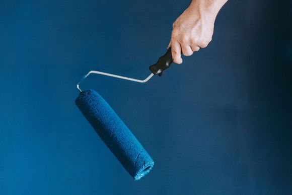 Person's hand holding a blue paint roller against a blue painted wall.