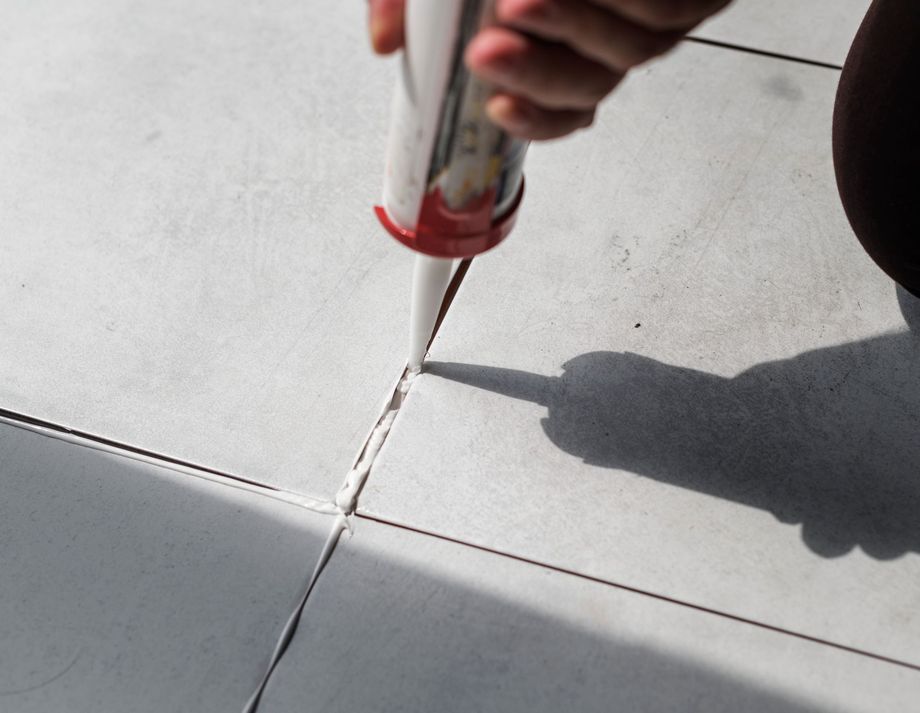 Person applying white caulk to the grout lines between white tiles on a floor.