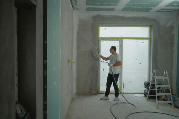 Man spraying drywall mud on a wall in a room under renovation.