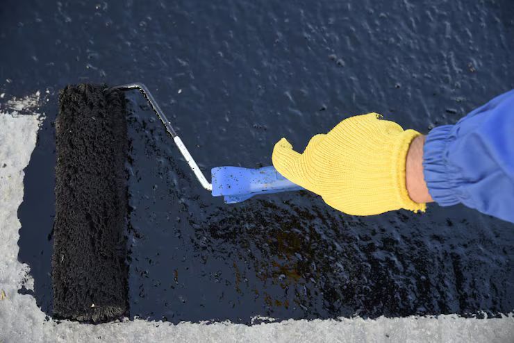 Hand with yellow glove painting black sealant on concrete with a roller.