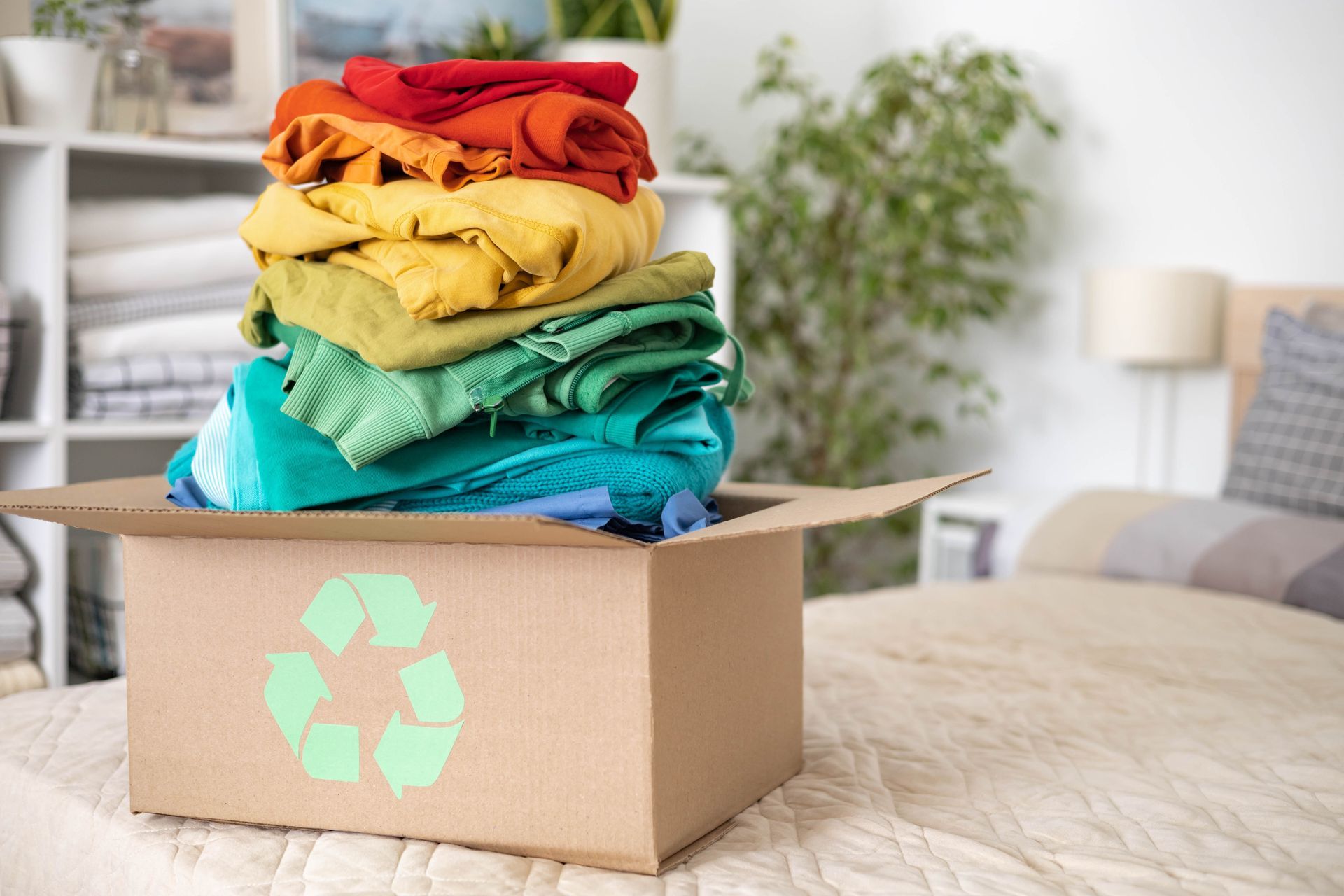 Cardboard box with a recycling symbol holds a colorful stack of clothing on a bed.