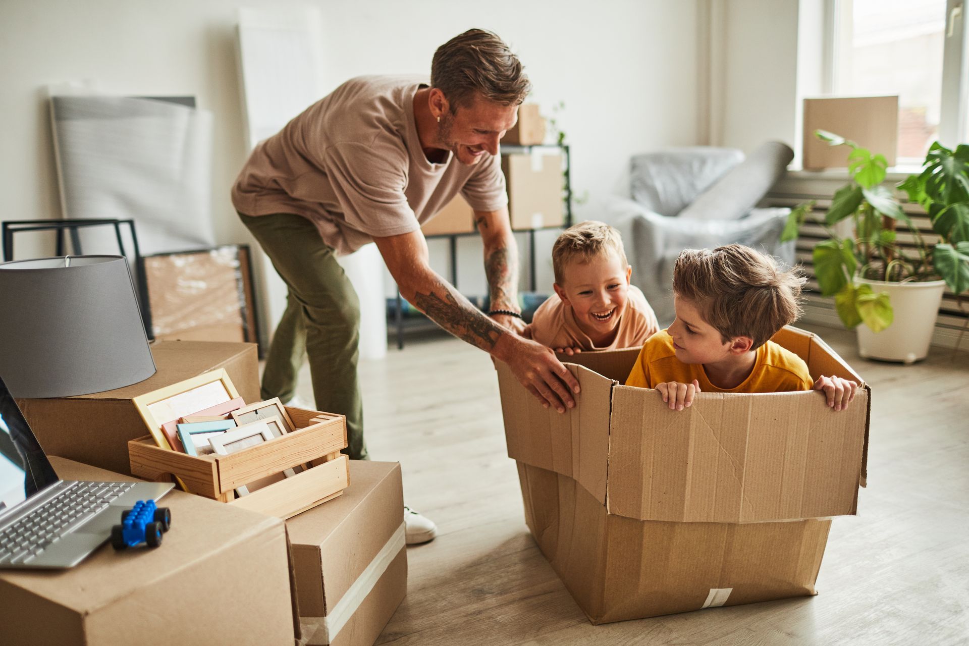 Man playing with two children in a cardboard box, surrounded by moving boxes in a room.