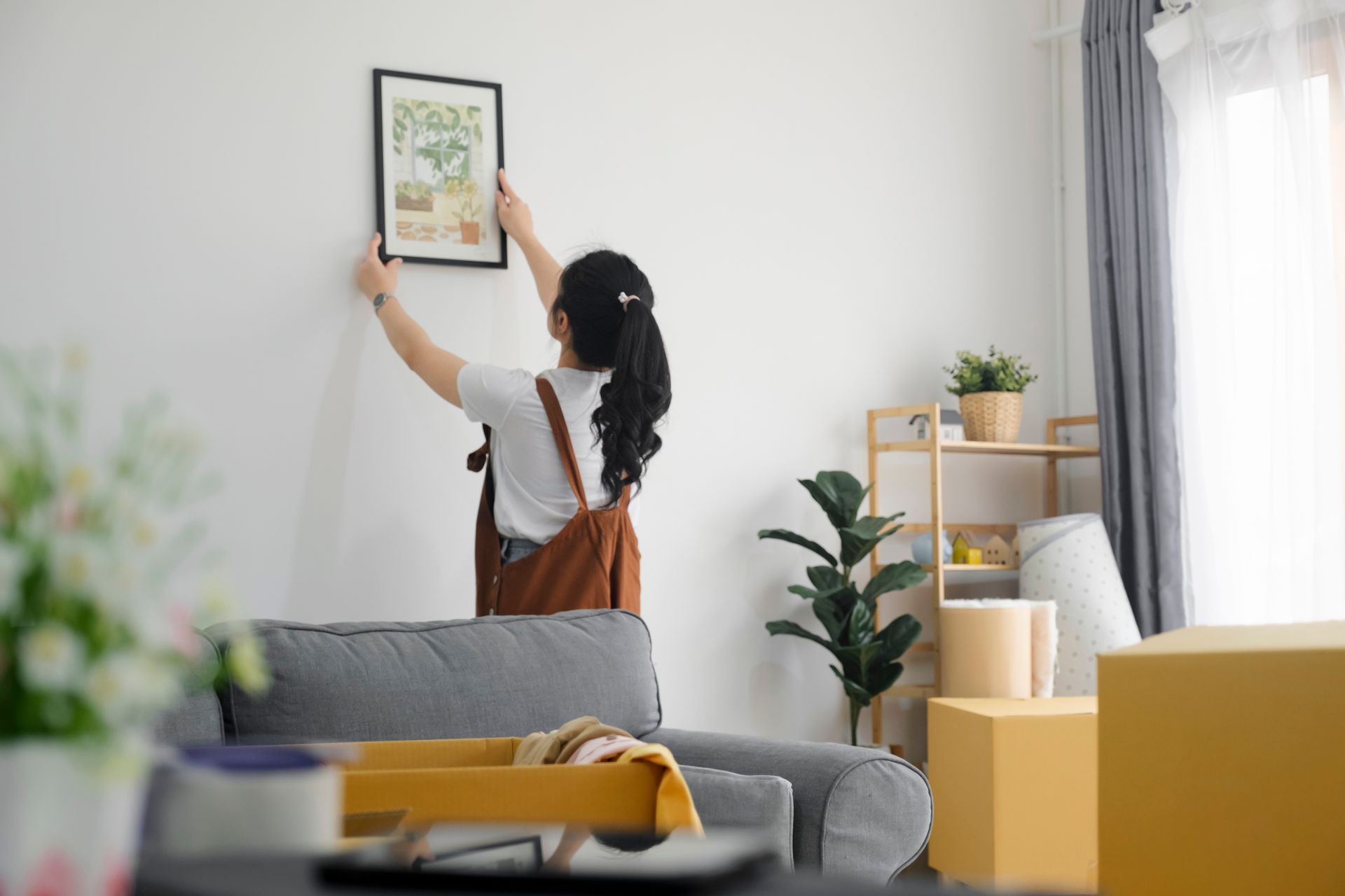 Person hangs framed art on a white wall in a bright living room with a gray sofa and plants.