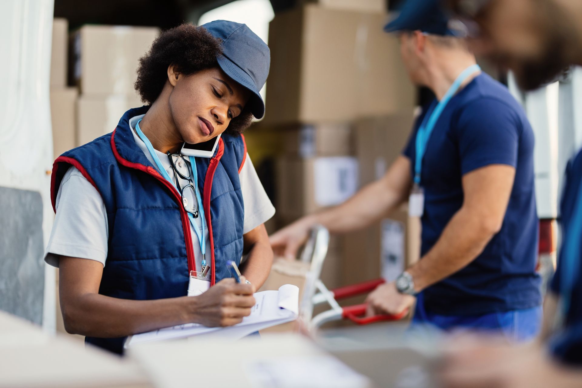 Woman in vest on phone, writing on clipboard near truck, with other workers and boxes.