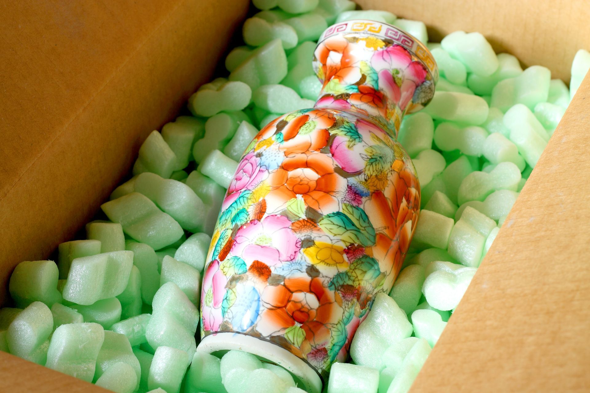 Colorful vase packed in a cardboard box surrounded by light green packing peanuts.