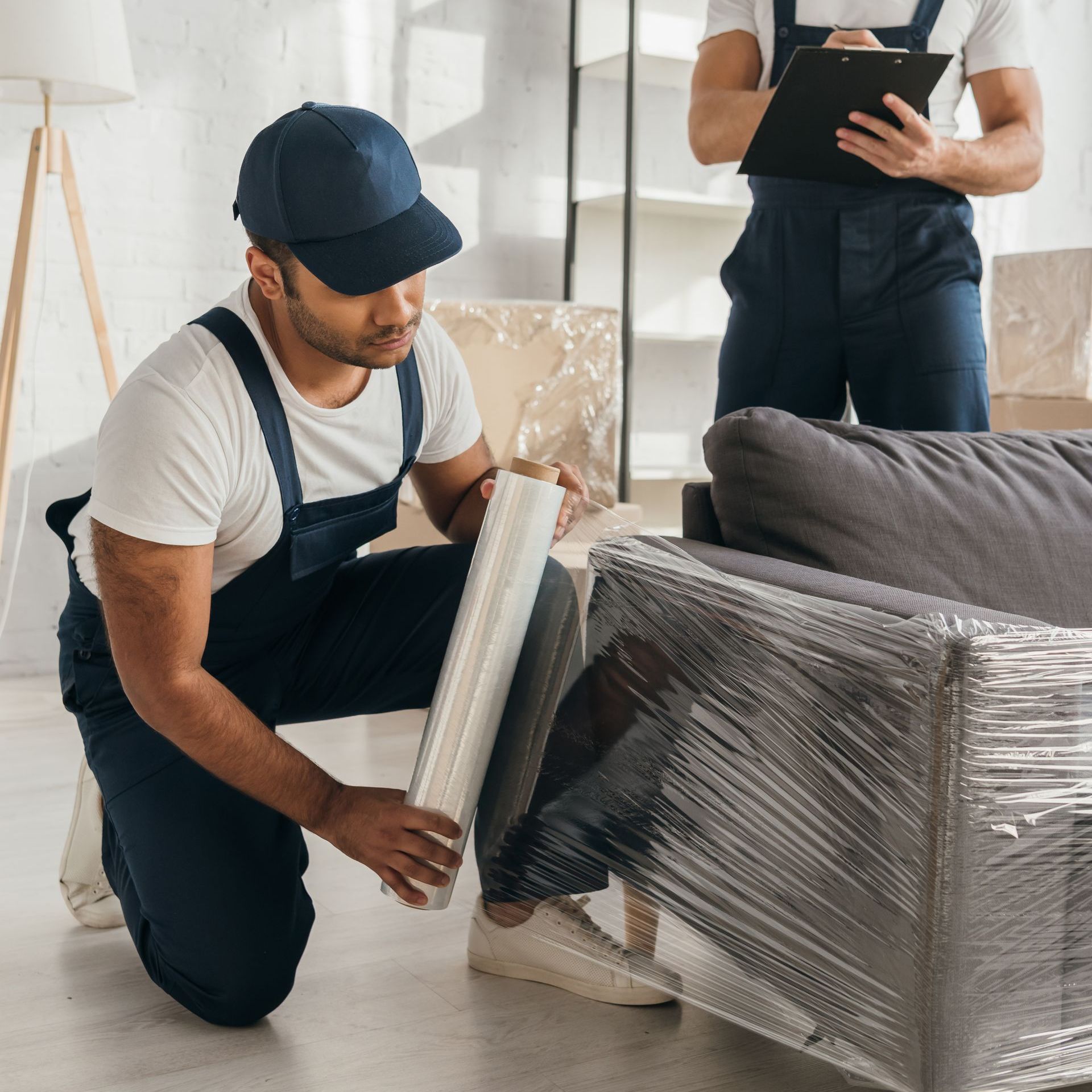 a man is wrapping a couch with plastic wrap while another man looks on .