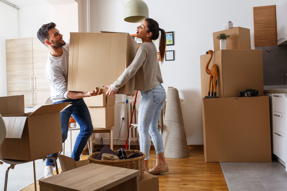 A man and a woman are carrying cardboard boxes in a living room.