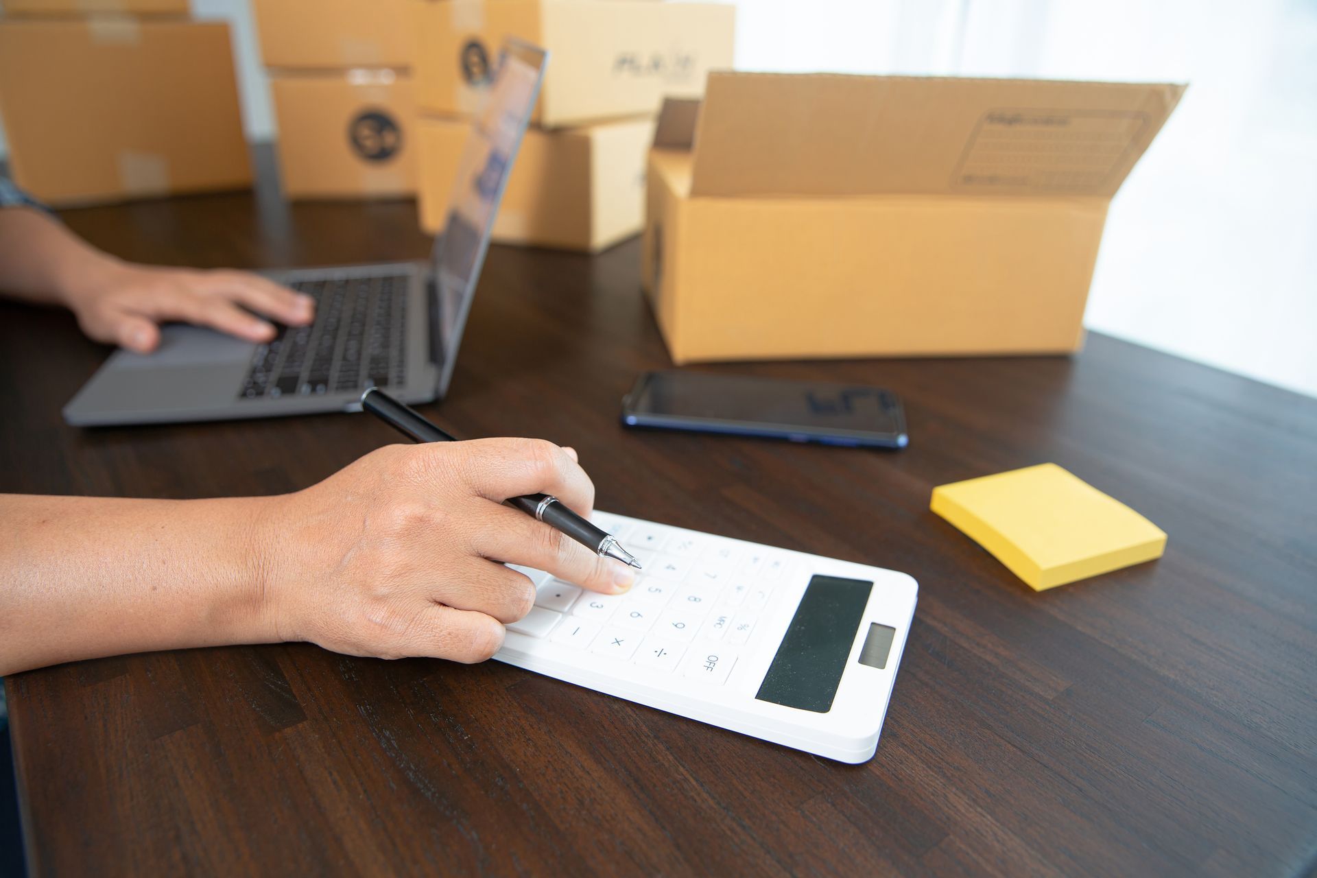 Person using a calculator with a laptop and boxes in the background.