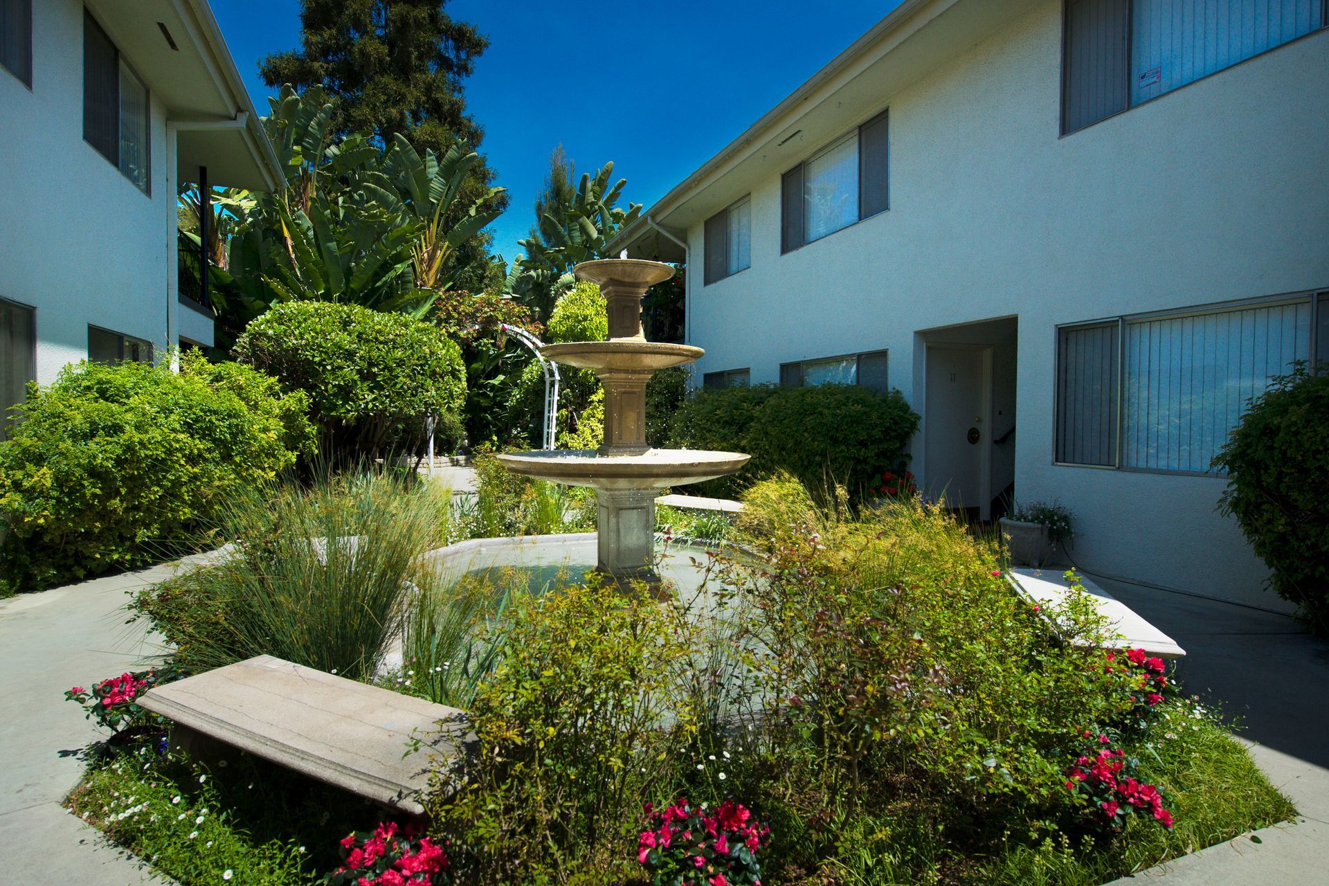A stone fountain sits in the center of a lush, landscaped courtyard between two white residential buildings.