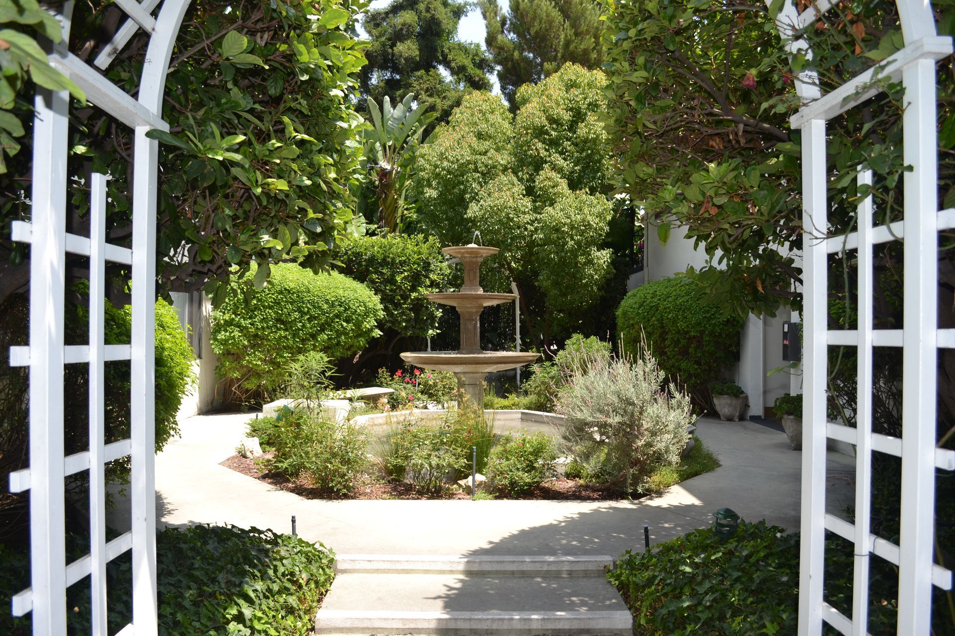 A white trellis frames a stone tiered fountain in a lush, green garden with paved paths.
