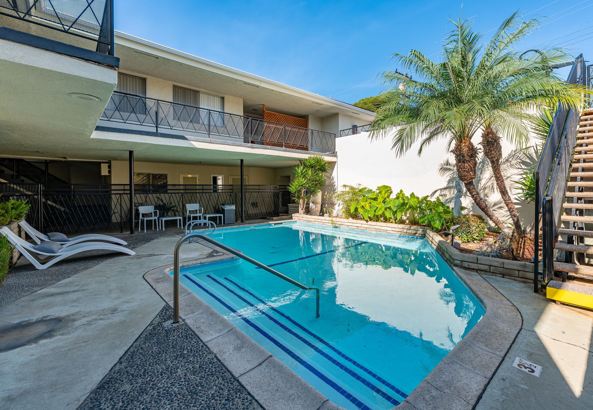 An outdoor swimming pool at a multi-story building, with stairs, palm trees, and patio furniture on a sunny day.
