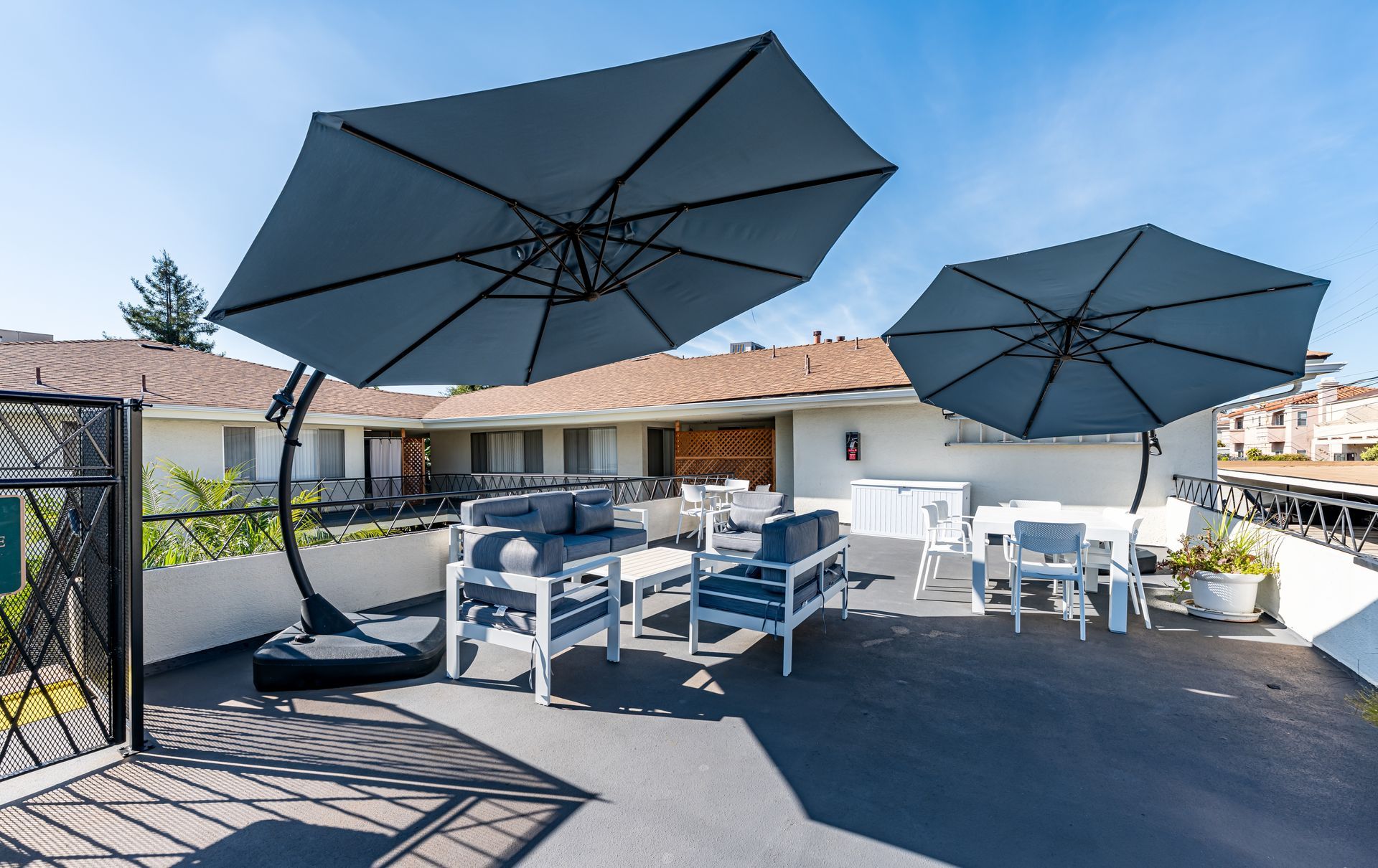 A sunny outdoor patio featuring lounge seating and dining tables shaded by two large, gray cantilever umbrellas.