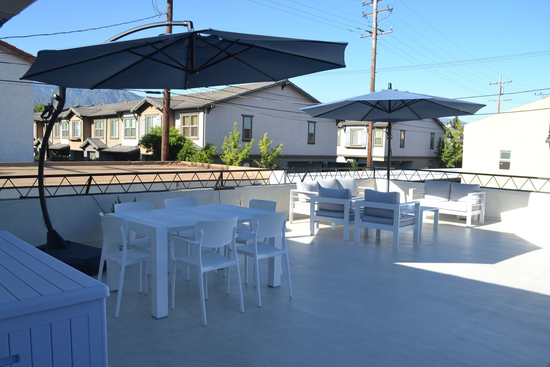 A sunlit rooftop patio with white furniture, tables, and large umbrellas set against a backdrop of suburban houses.