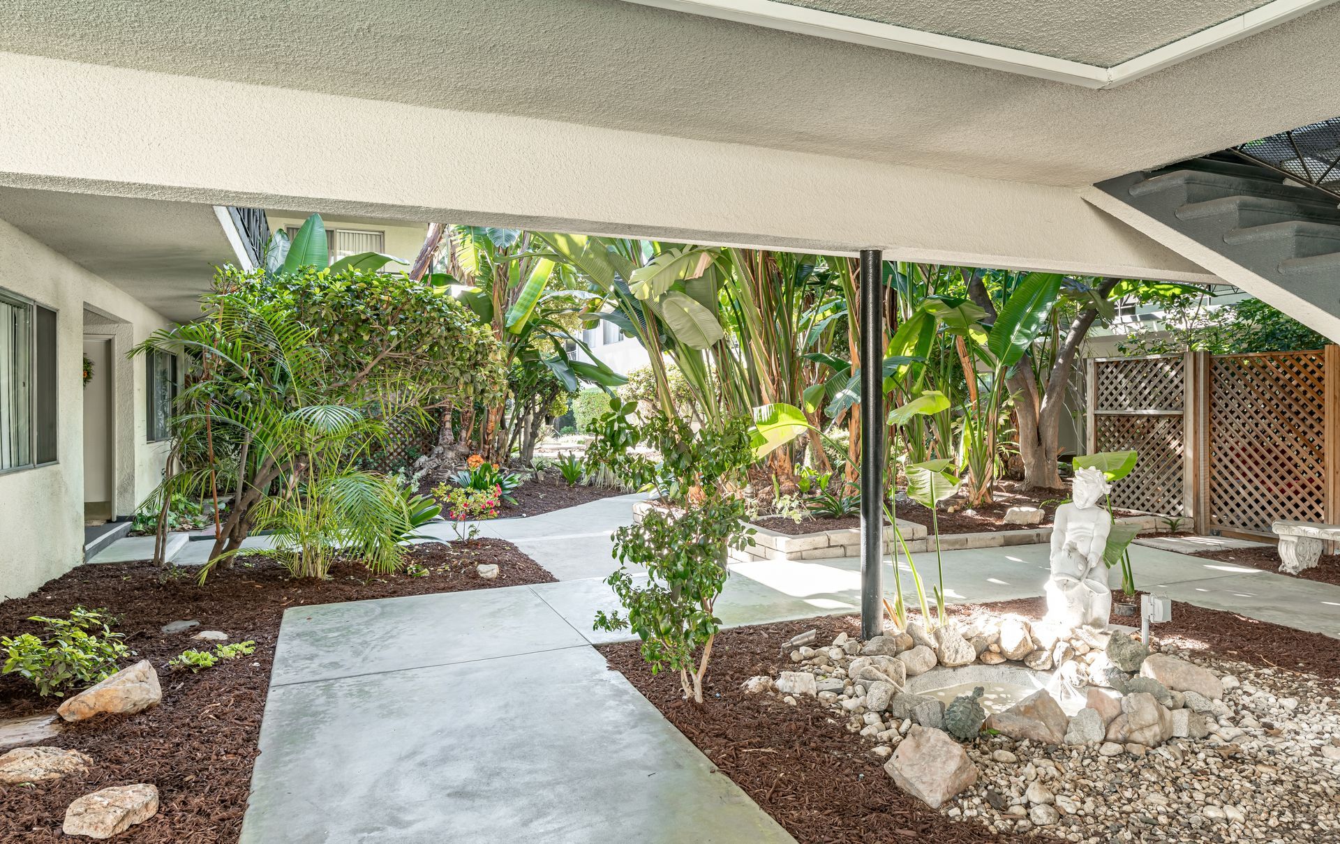 A covered walkway leads through a landscaped outdoor area with lush tropical plants, rocks, and a small stone statue.