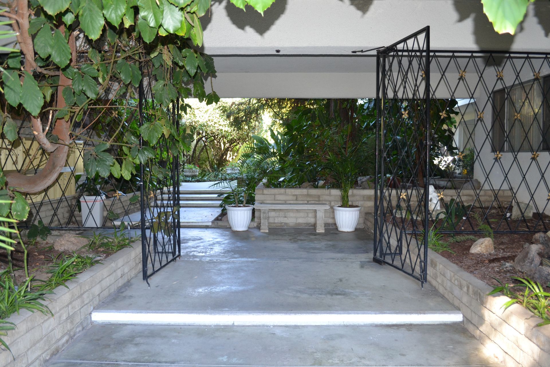 An open wrought-iron gate leads to a concrete patio with a stone bench and potted plants under a covered entryway.