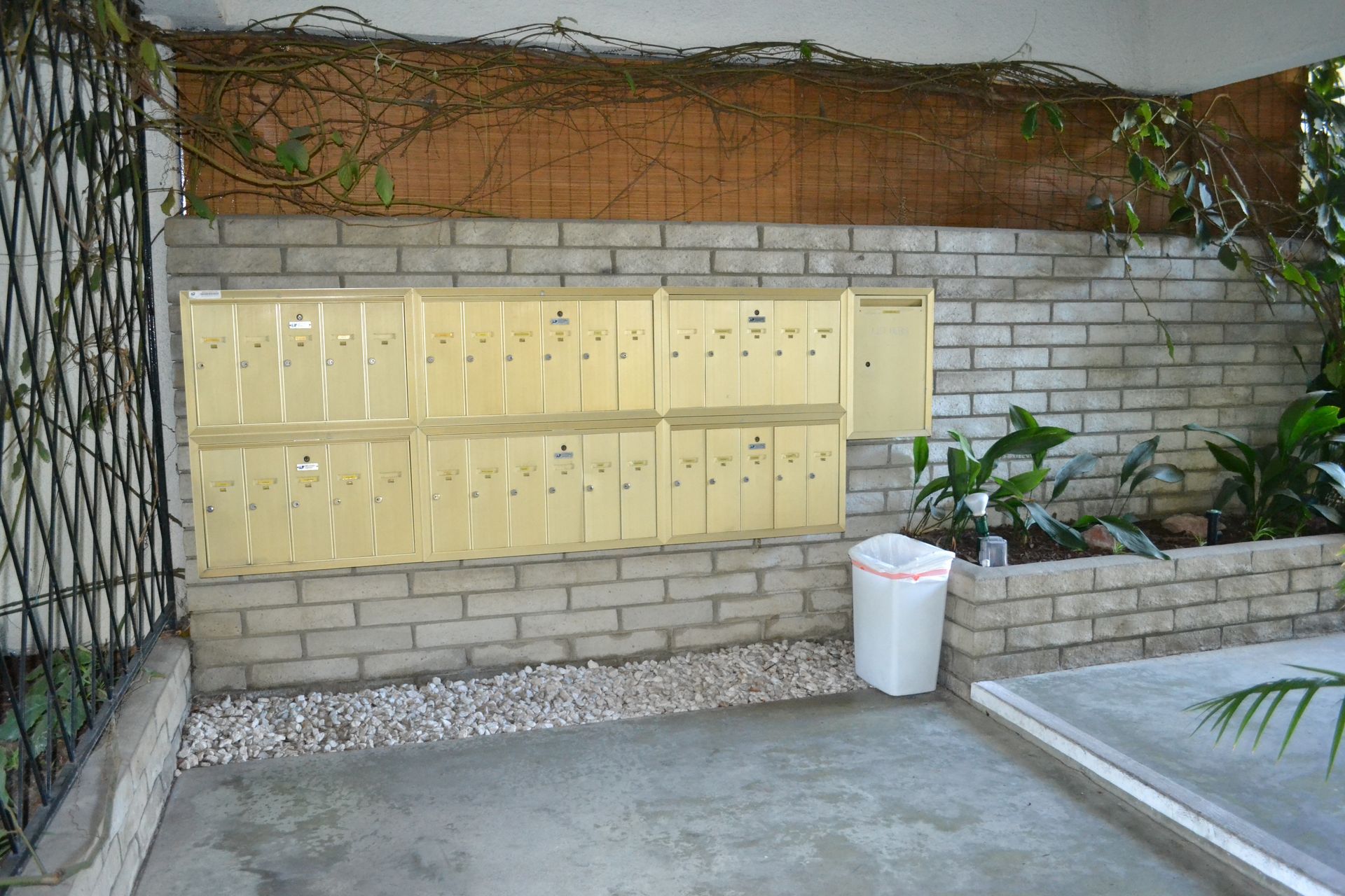 A wall of gold-colored mailboxes mounted on a brick wall next to a white trash bin in an outdoor courtyard.