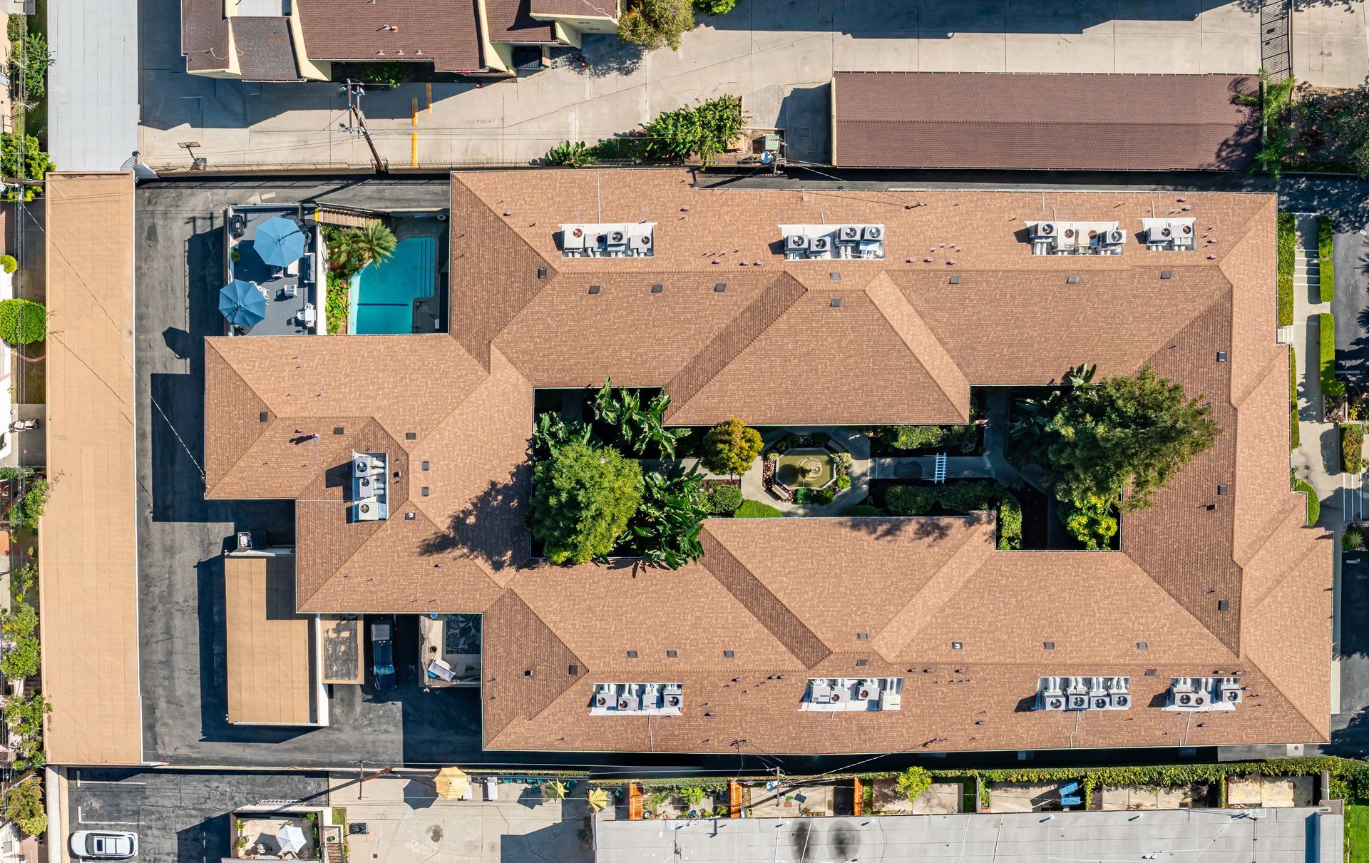 A top-down aerial view of a U-shaped apartment building with a brown shingled roof, a central courtyard, and a swimming pool.