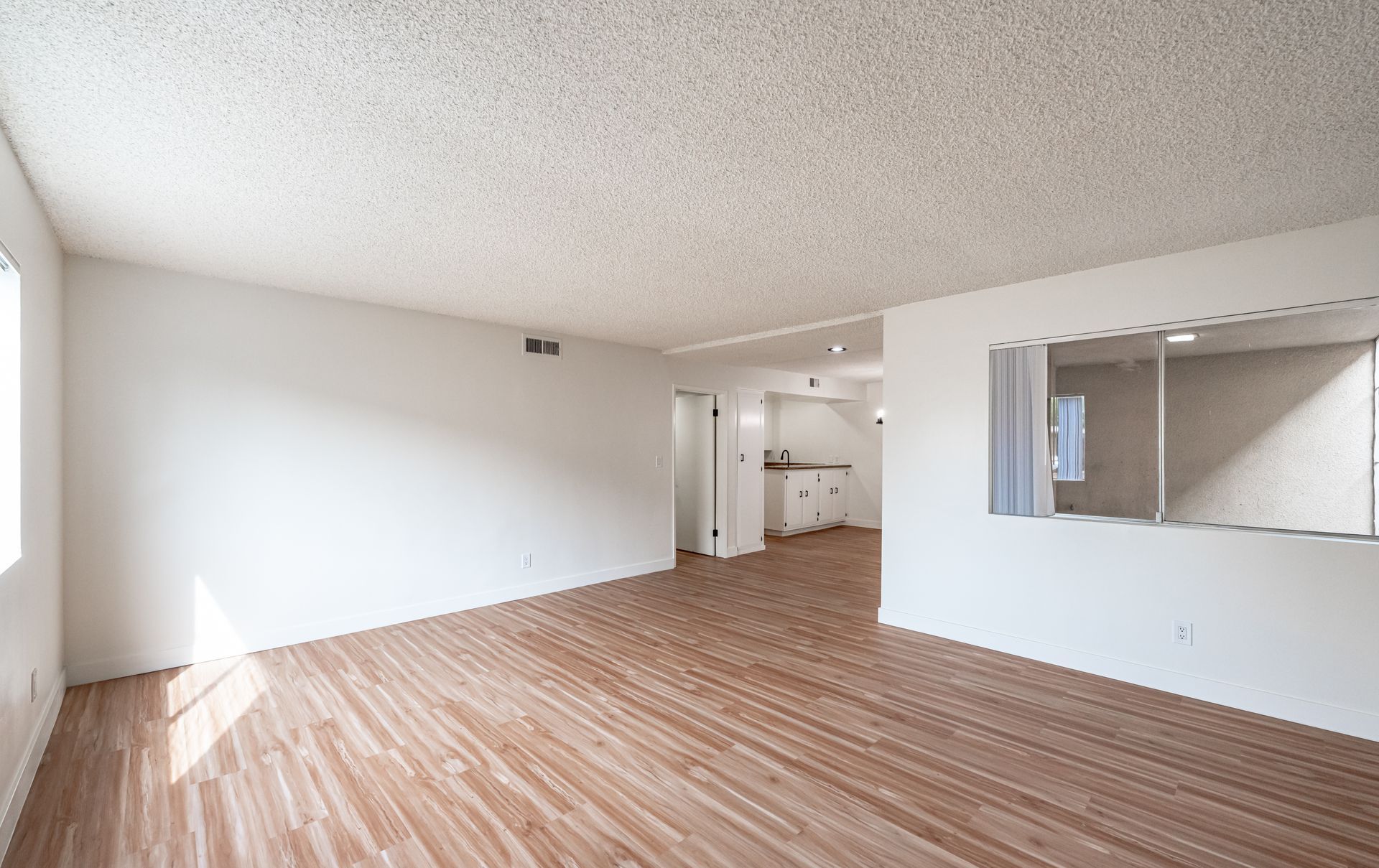 An open-concept living area with light wood-style flooring, white walls, a textured ceiling, and a kitchen pass-through.