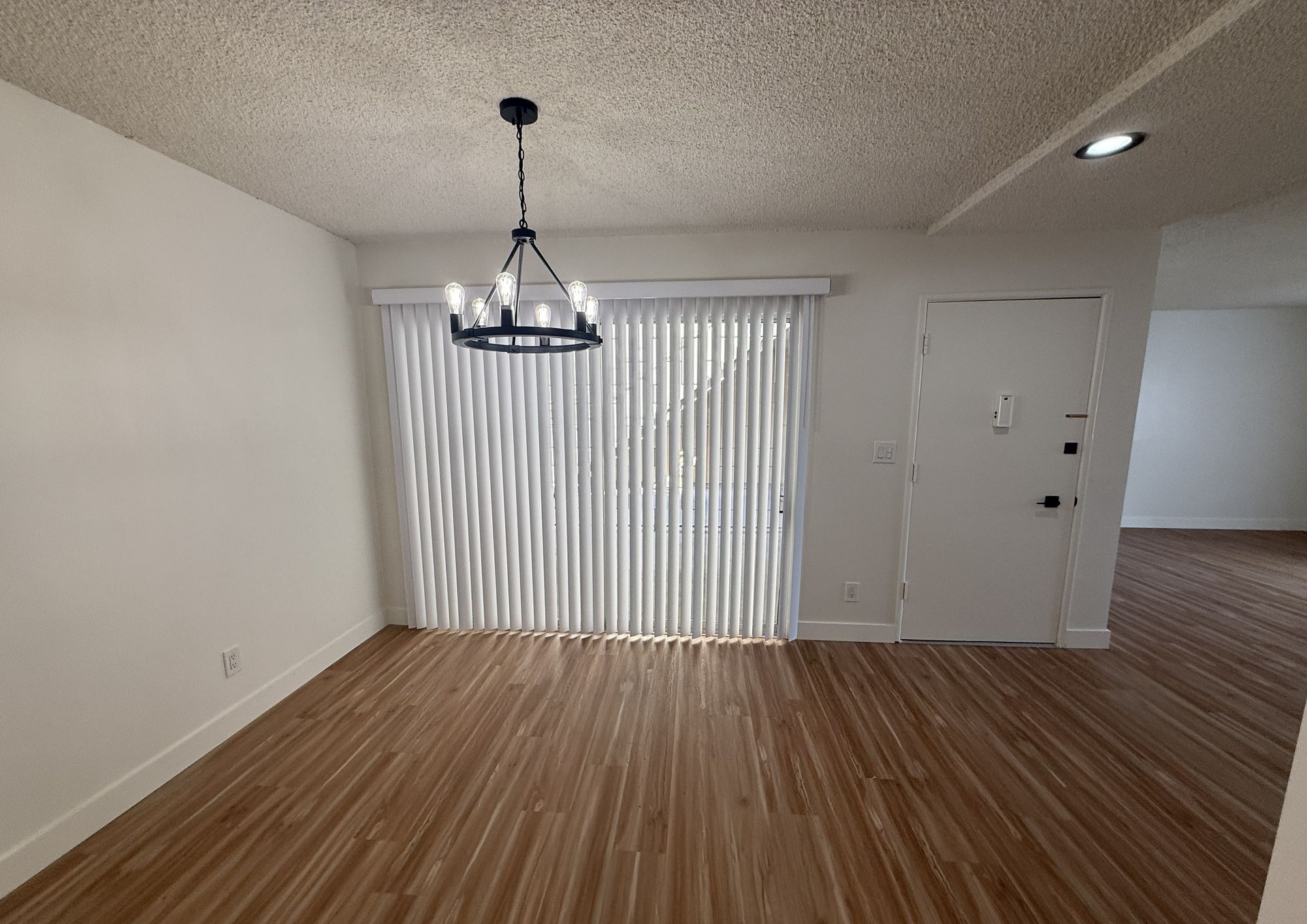 A dining area with wood-look flooring, white walls, a black chandelier, vertical blinds on sliding doors, and a white door.
