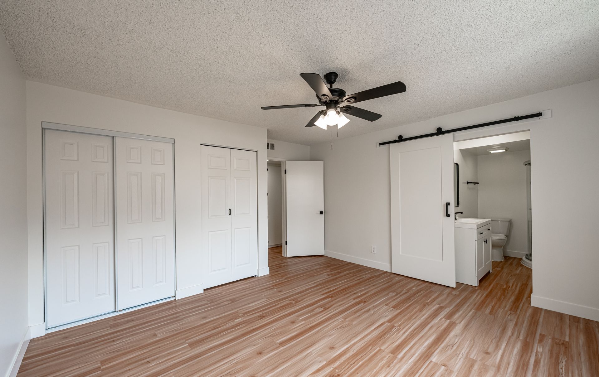 An unfurnished bedroom with white walls, light wood-look floors, a ceiling fan, sliding closet doors, and a bathroom.