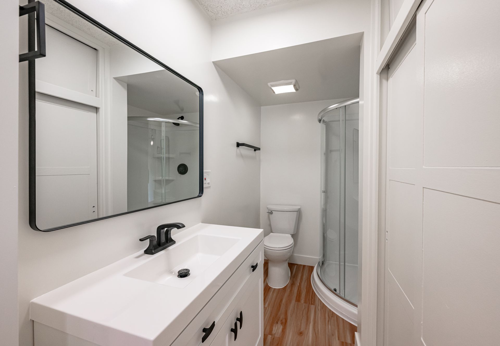 Modern bathroom with a white vanity, black-framed mirror, toilet, and a glass-enclosed corner shower on wood floors.