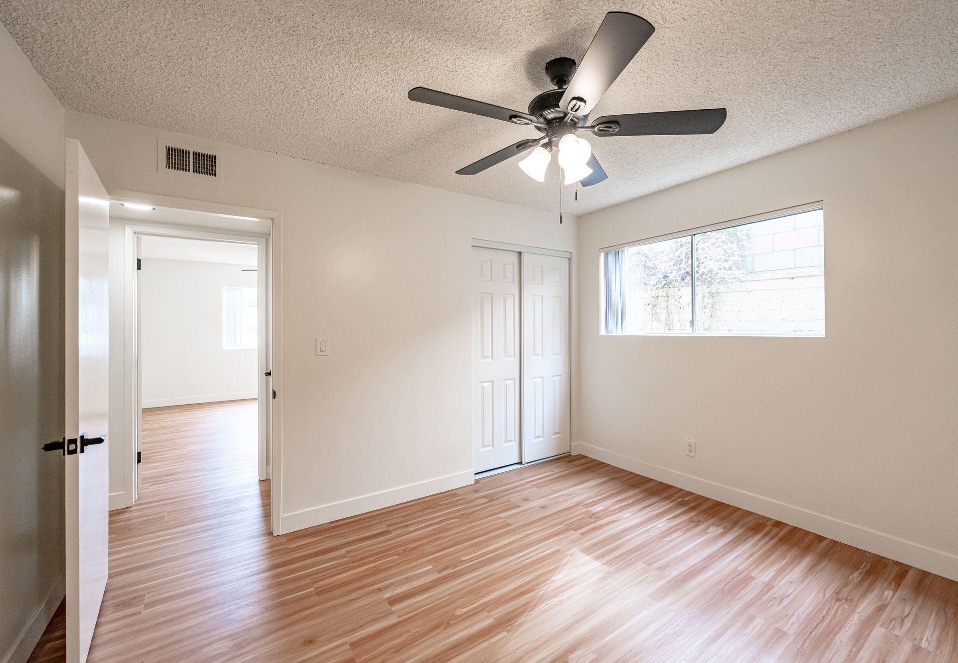 An empty, neutral-colored bedroom featuring wood-style flooring, a ceiling fan, a closet, and an open doorway.