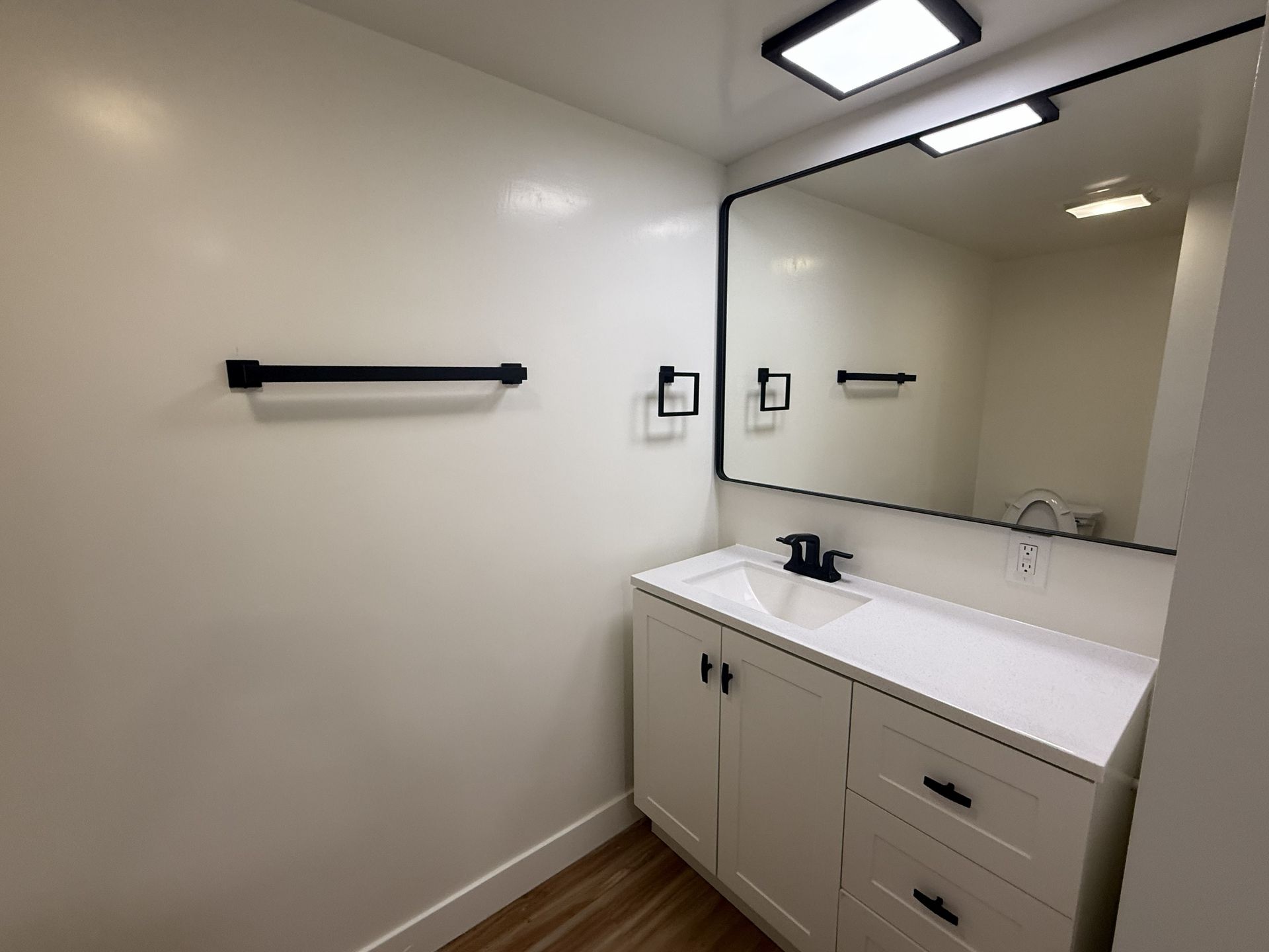 A modern bathroom vanity with a white cabinet, black hardware, a rectangular mirror, and a towel bar on a white wall.