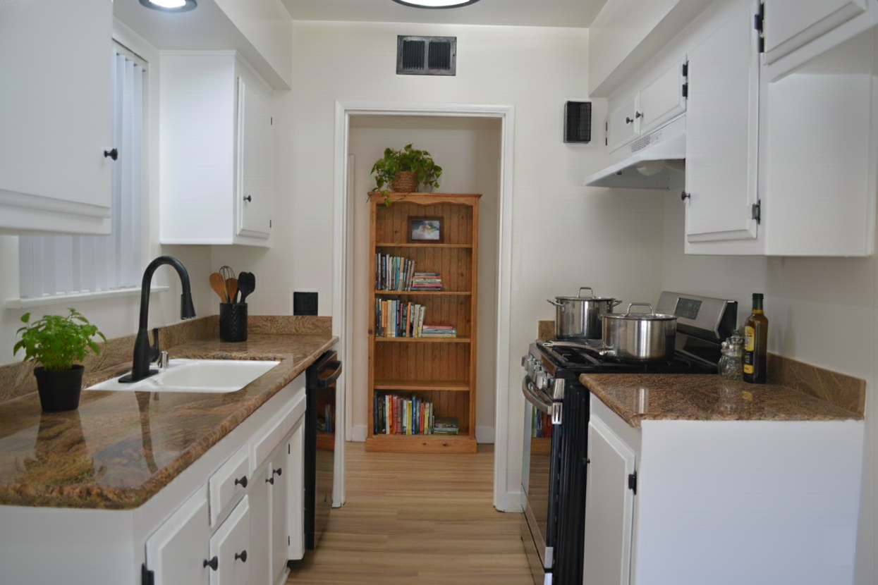 A narrow galley kitchen with white cabinets, brown granite countertops, black appliances, and a wood bookshelf in the back.
