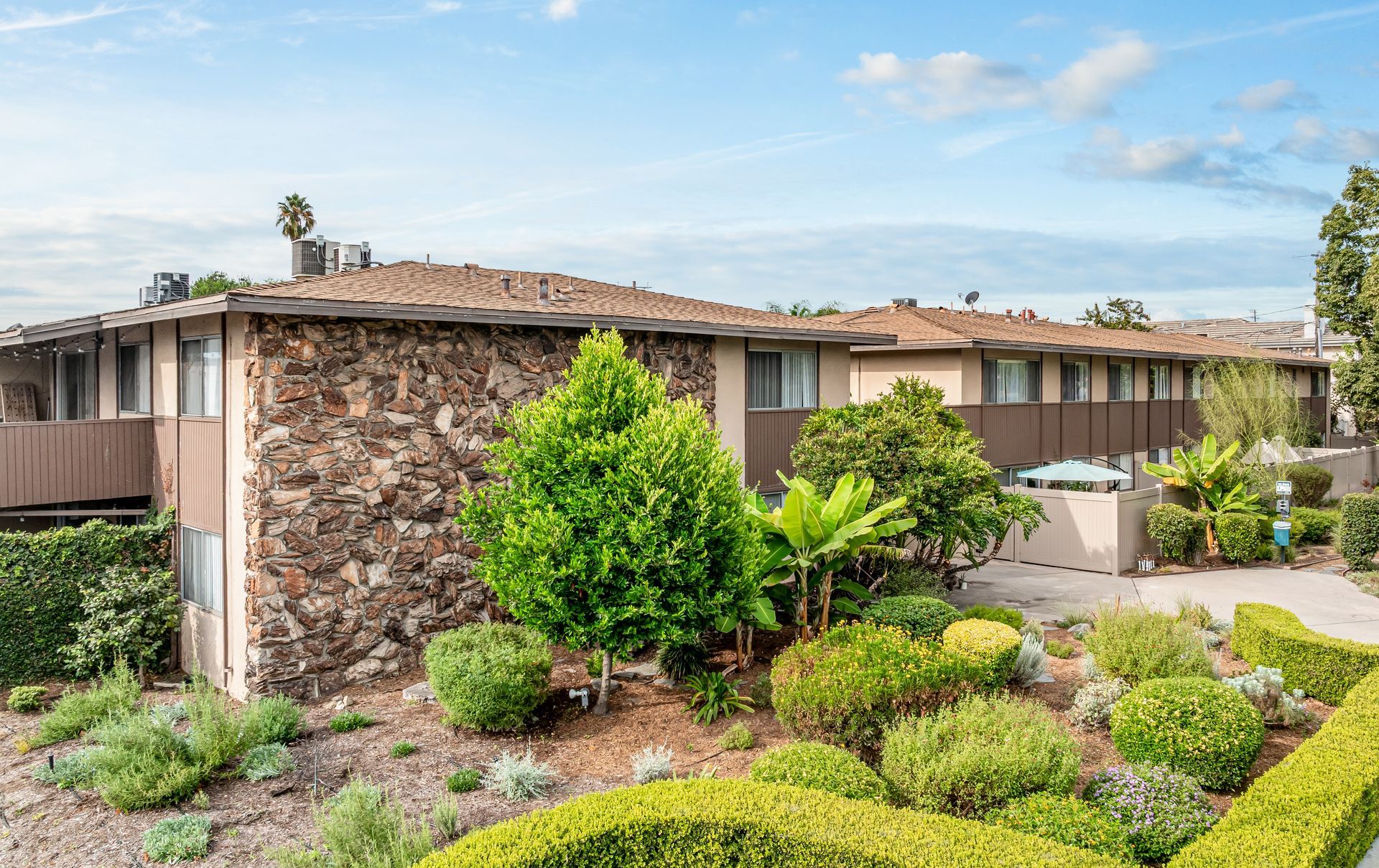 A two-story apartment building with stone siding and brown trim, surrounded by landscaped bushes and greenery.