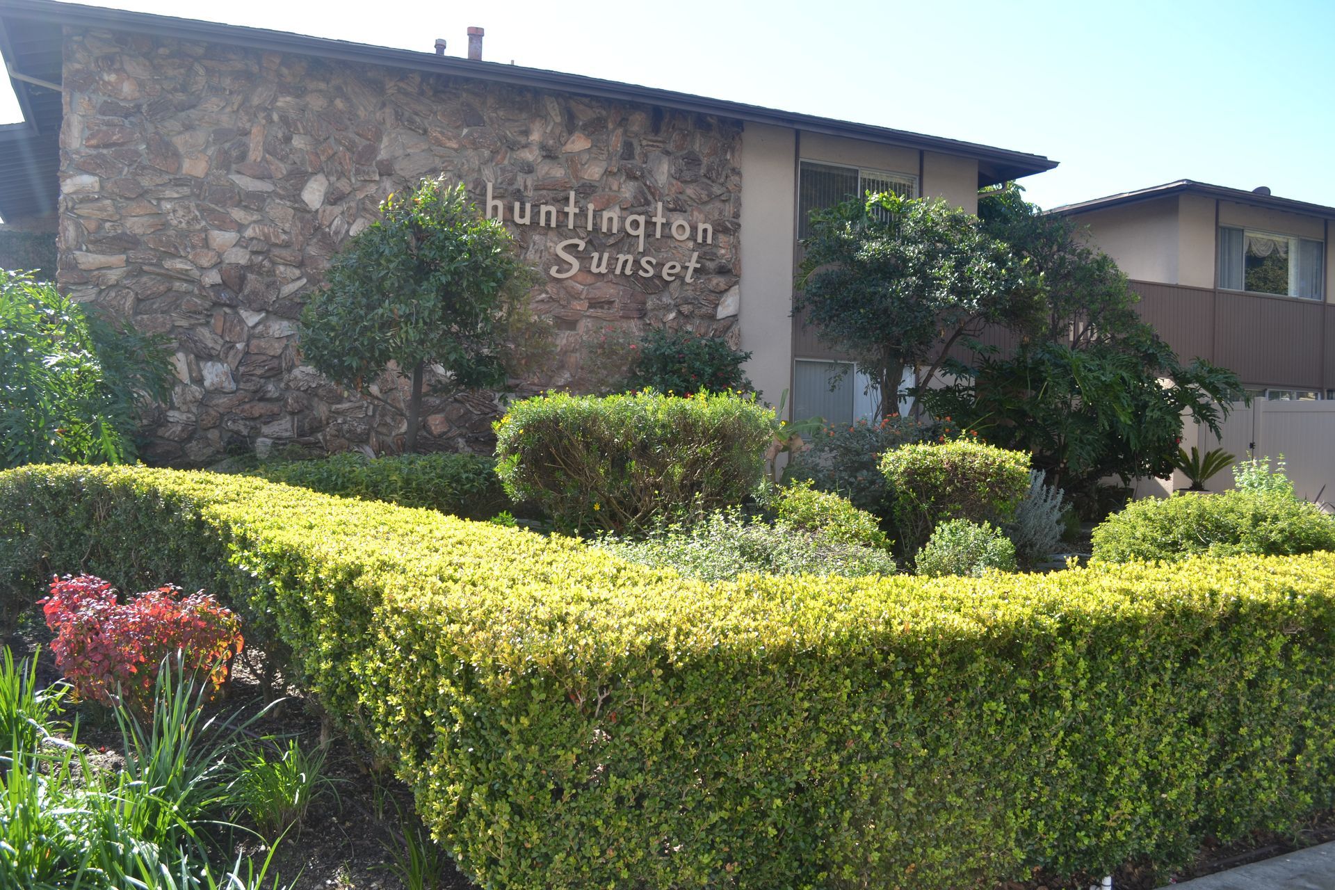 A two-story apartment complex with a stone facade, trimmed green hedges, and trees under a clear blue sky.