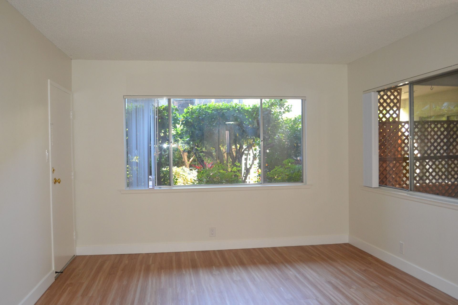 An empty room with light wood floors, beige walls, and two windows looking out onto greenery and a lattice screen.