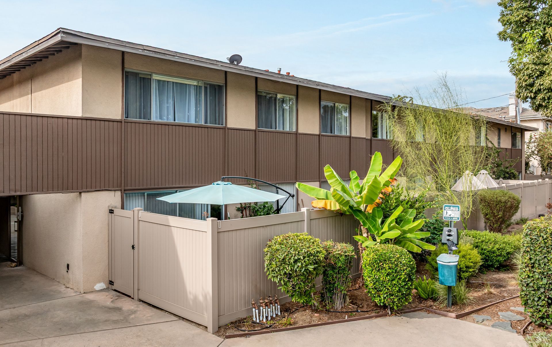 A two-story apartment complex with beige stucco walls, brown trim, and a small fenced-in patio area with a green umbrella.