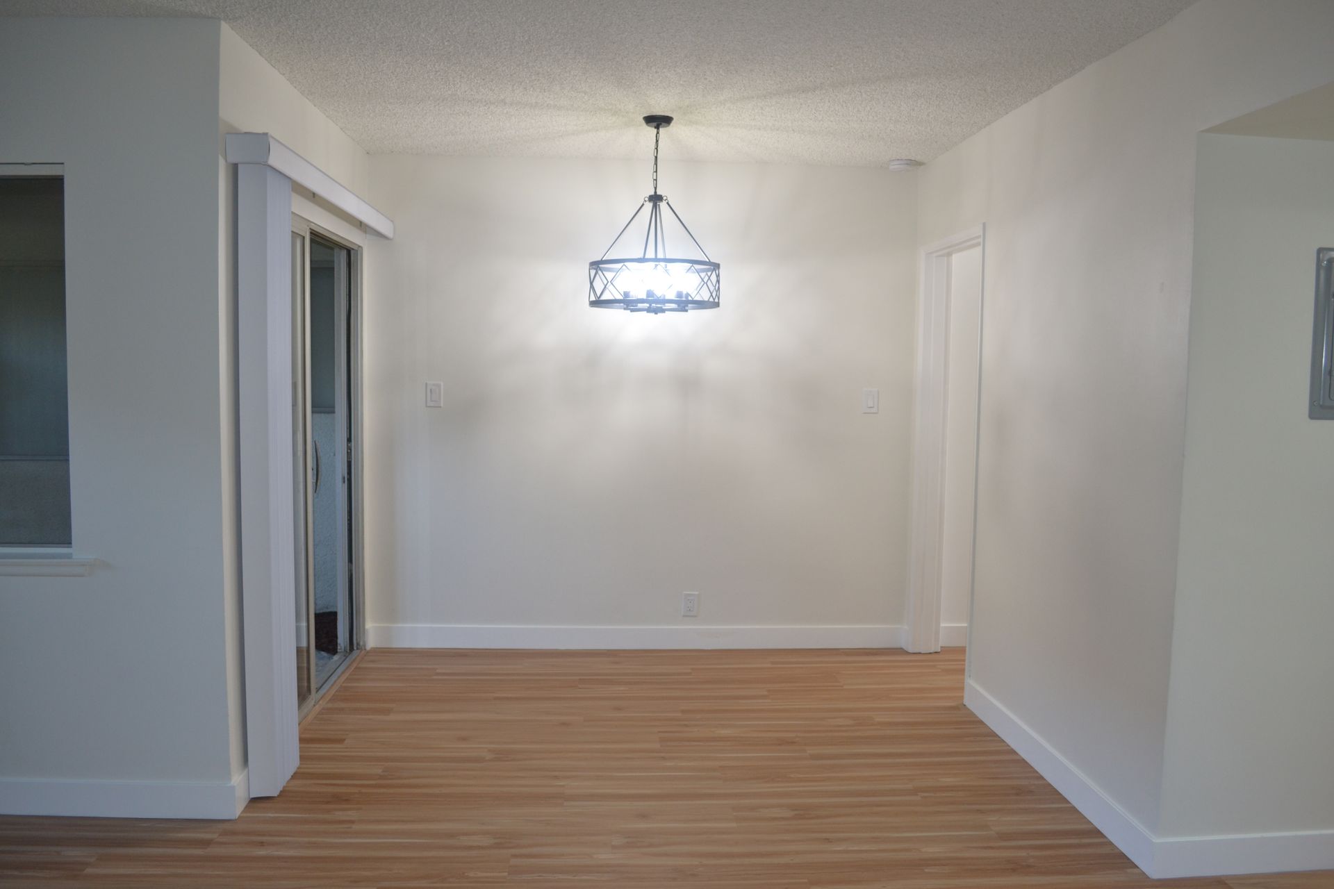 An empty dining area with light hardwood floors, beige walls, white trim, and a modern chandelier hanging centered.
