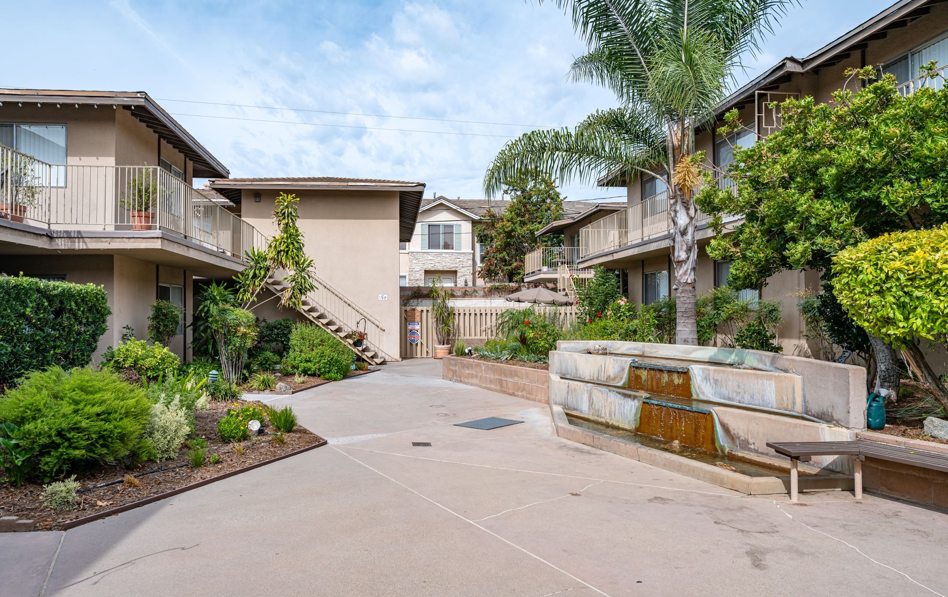 A paved courtyard in an apartment complex with tan buildings, lush landscaping, a stone fountain, and a palm tree.
