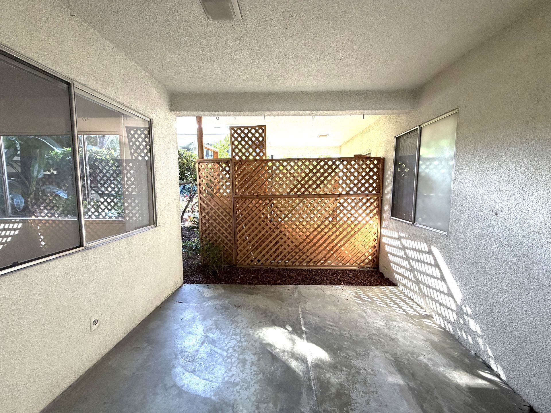 A covered patio with light-colored stucco walls, two windows, and a wood lattice privacy screen at the back.