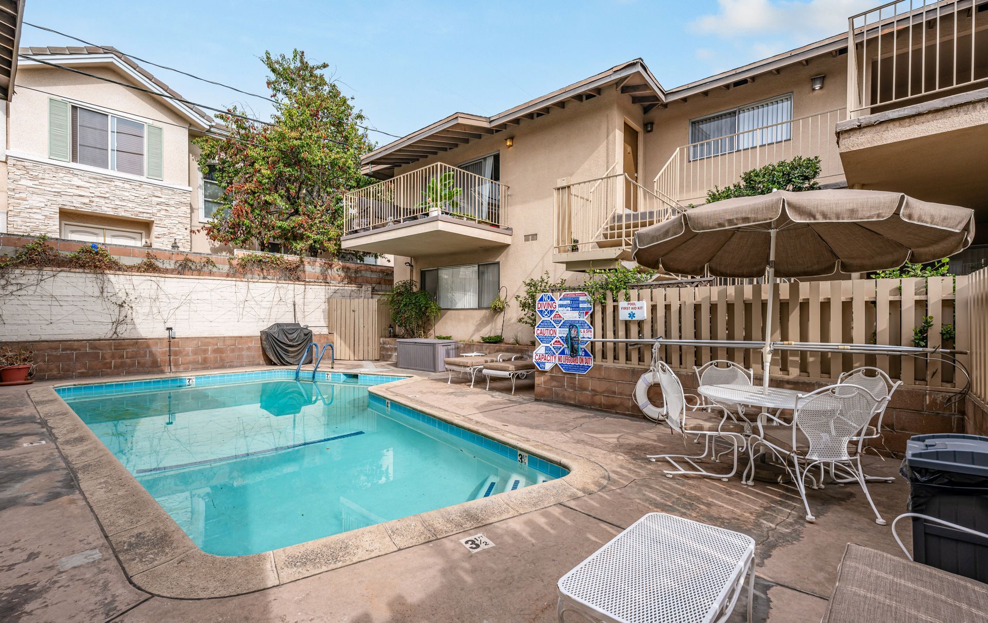 A beige two-story building overlooks a rectangular backyard swimming pool with a patio set and umbrella on the deck.